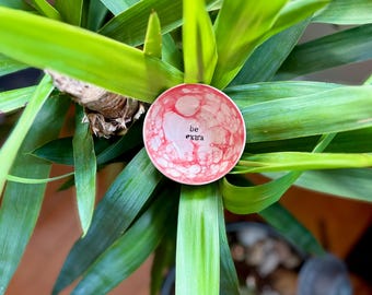 May include: A small, pink and white ceramic dish with a bubbled glaze. The dish has the words "be extra" written on it in black.