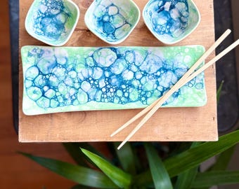 May include: A green and blue ceramic serving dish with three matching small bowls and a pair of wooden chopsticks. The dish and bowls have a bubble-like pattern.