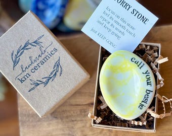 May include: A small, yellow and white ceramic worry stone with the text "call your bestie" on it. The stone is in a small box with brown paper shredding. The box has a label that says "handmade km ceramics". A separate card with the text "worry stone" and instructions on how to use the stone is also included.