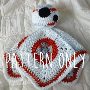 May include: A handmade crochet baby toy in the shape of a BB-8 droid. The toy is white with orange and gray accents, and a black eye. The text "PATTERN ONLY" is across the bottom of the toy.