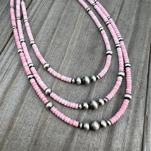 Könnte beinhalten: Eine mehrreihige Halskette mit drei Strängen aus rosa Muschelkugeln und silberfarbenen Metallperlen. Die Halskette wird auf einer verwitterten Holzoberfläche präsentiert, die ihre natürlichen Töne und Texturen hervorhebt.