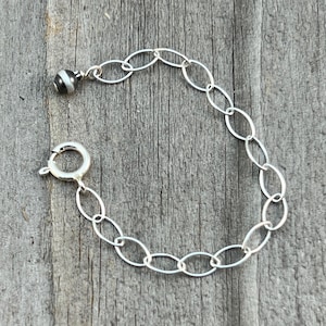 May include: A silver-colored chain extender with an oval link design. One end features a small, round charm with black and white stripes, while the other has a spring ring clasp. The chain is displayed on a weathered wooden surface.