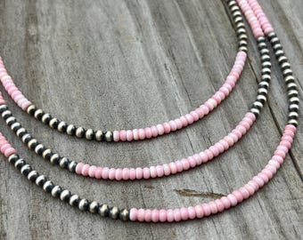 May include: Three-strand necklace featuring pink beads and silver-colored metal beads. The necklaces are displayed on a weathered wooden surface, showcasing the jewelry's design and color contrast.