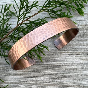 May include: A hammered copper cuff bracelet with a smooth, polished finish. The bracelet is made of a single piece of copper that has been hammered to create a textured surface.