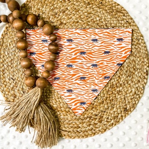 May include: A triangular dog bandana with a white background and an orange zebra stripe pattern. Small navy blue panther silhouettes are scattered across the fabric. The bandana is displayed on a woven mat with wooden beads.