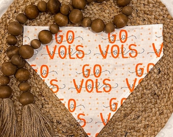 Könnte beinhalten: Ein weißes Bandana mit orangefarbenem Text, der "Go Vols" wiederholt auf weißem Hintergrund mit orangefarbenen Punkten. Das Bandana liegt auf einer braunen gewebten Matte mit einem braunen Quasten.