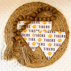 May include: Three white fabric dog collars with paw prints and the word "Tigers" repeated in purple and yellow lettering. The collars are lying on a woven straw mat with a brown and beige beaded garland.