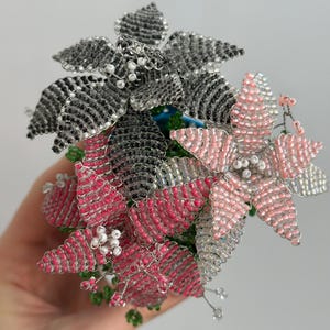 May include: Close-up of beaded floral arrangement. The arrangement features three flowers in shades of pink, silver, and black. The petals are made of beads, and the stems are wire. The flowers are clustered together, creating a decorative display.