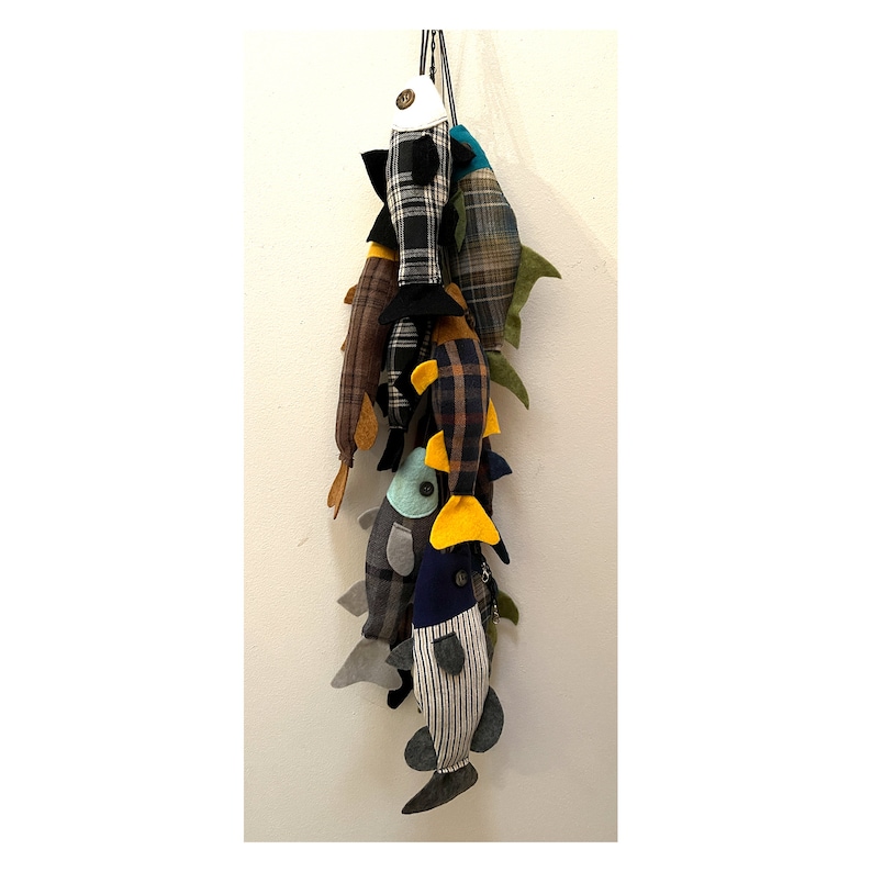 May include: A group of handmade fabric fish toys hanging from a string. The fish are made from different patterned fabrics, including plaid, striped, and solid colors. The fish have yellow, blue, and green accents.