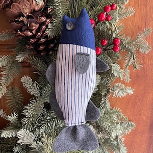 May include: A handmade blue and white striped fish ornament with a gray felt tail and a blue button eye. The ornament is hanging from a green pine branch.
