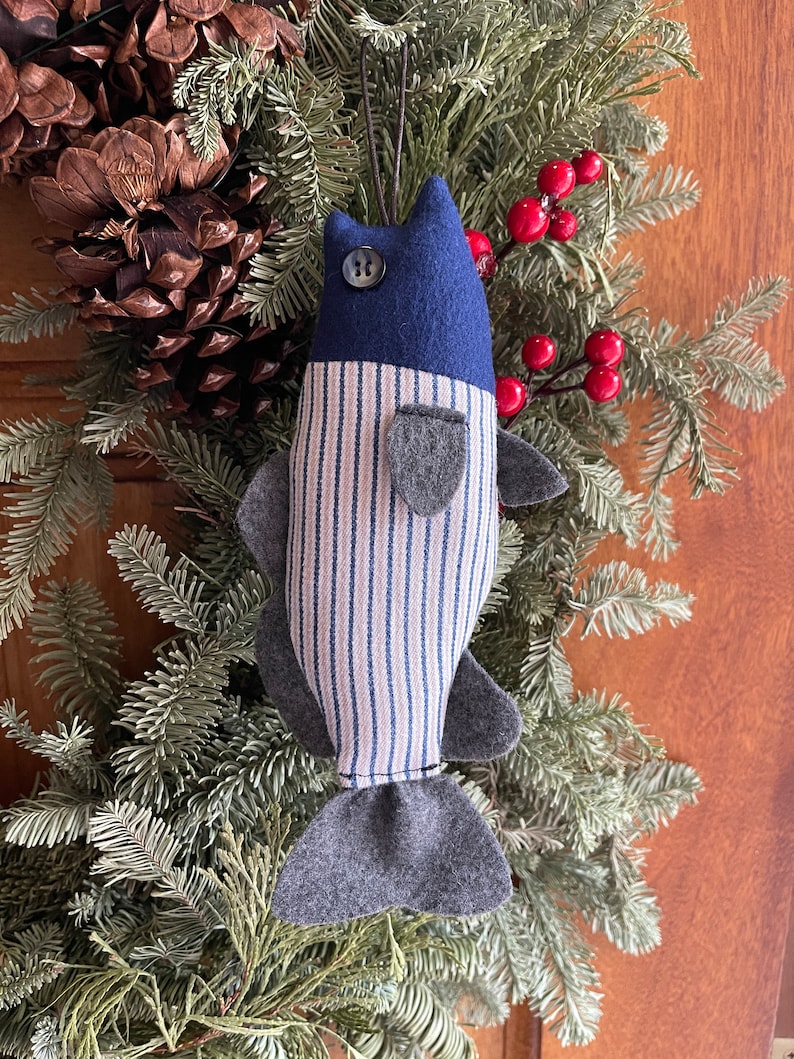 May include: A handmade blue and white striped fish ornament with a gray felt tail and a blue button eye. The ornament is hanging from a green pine branch.