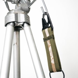 Könnte beinhalten: Ein silbernes Stativ mit einem schwarzen und grünen Gurt. Der Gurt hat ein digitales Tarnmuster und einen schwarzen Metallclip und -ring. Das Stativ ist aus Metall und hat drei Beine.