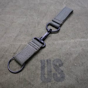 Könnte beinhalten: Ein olivgrüner Canvas-Gurt mit einem schwarzen Metallverschluss und Ringen. Der Gurt ist mit einem passenden olivgrünen Canvas-Hintergrund mit den Buchstaben "US" bedruckt.