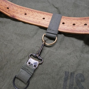 May include: Close-up of a military-style canvas bag with a leather belt and metal hardware. The belt is light brown with a decorative pattern. A green canvas strap connects to a metal clasp and a ring. The bag is olive green.