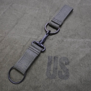 May include: An olive green canvas strap with a metal clasp and two metal rings. The strap is connected to a larger olive green canvas surface with the letters "US" printed on it.