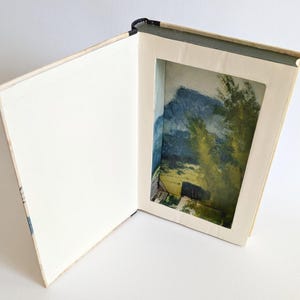Könnte beinhalten: Ein offenes Buch mit einem Landschaftsgemälde, das in einem ausgeschnittenen Bereich sichtbar ist. Das Gemälde zeigt eine Bergkette, Bäume und ein Gebäude, dargestellt in Blau-, Grün- und Gelbtönen. Die Seiten des Buches sind weiß, und der Einband ist hellbeige.