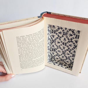 Peut inclure: Un livre évidé avec un compartiment rectangulaire découpé dans les pages. Le côté gauche affiche du texte imprimé en allemand, tandis que le côté droit révèle un intérieur caché doublé d'un tissu à motifs noir et blanc avec des petits éventails.