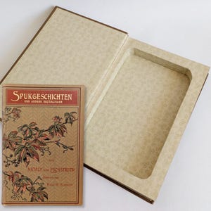 Könnte beinhalten: Eine offene, vintage Buchbox mit einem Buch mit dem Titel "Spukgeschichten" daneben. Die Box ist beige mit einem floralen Muster, das Buch hat einen braunen Einband mit rotem Text und botanischen Illustrationen.