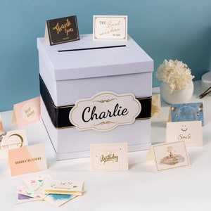 May include: White card box with a black ribbon and a gold label that says "Charlie". The box is surrounded by various cards with different messages, including "Happy Birthday", "Best Wishes", "Thank You", "Congratulations", "Smile", and "Dreams".