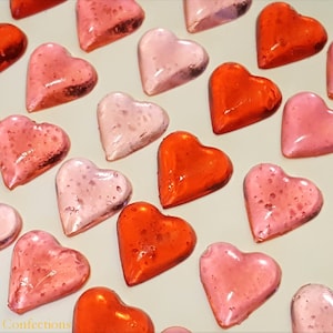 May include: Assortment of heart-shaped candies in red, pink, and light pink. The candies are arranged in rows on a white surface.