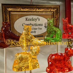 May include: A variety of colorful handmade candy lollipops shaped like bears and cats. The lollipops are displayed on sticks and are arranged in a row. The lollipops are made with clear candy and have a variety of colors, including red, green, blue, and yellow. The lollipops are displayed on a white surface. The sign above the lollipops reads "Keeley's Confections Clear Toy Candy".