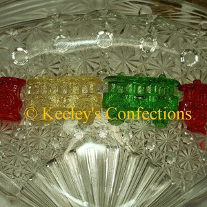 May include: Four colorful, transparent train car candies in red, yellow, green, and red. The candies are arranged on a clear, patterned glass dish. The text "Keeley's Confections" is visible.