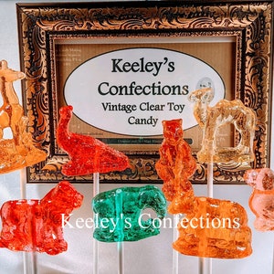 May include: A collection of vintage clear toy candy animal lollipops in various colors, including orange, red, and green. The lollipops are displayed in front of a framed sign that reads "Keeley's Confections Vintage Clear Toy Candy."