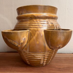May include: A brown ceramic planter with a textured, glazed finish. The pot has a rounded base, a central section with two side pockets, and a wide top. Horizontal ridges decorate the lower part of the planter.