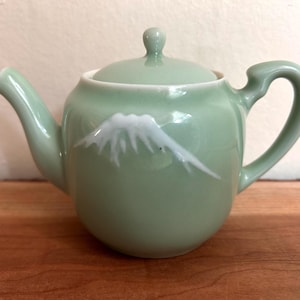 May include: A light celadon green ceramic teapot with a matching lid, a curved handle, and a spout. The body of the teapot features a white, stylised graphic of a mountain peak with snow-capped ridges centred on the front surface.