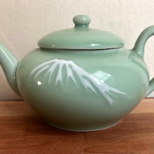 May include: A light sage green ceramic teapot with a matching lid, a curved handle, and a spout. The body of the teapot features a white graphic design depicting a mountain peak with snow-capped ridges, rendered in a smooth, glossy finish.