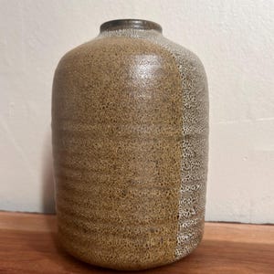 May include: A brown ceramic vase with a textured surface. The vase has a rounded body and a narrow neck. The color is a mix of brown and tan, with a speckled pattern on one side. The vase is sitting on a wooden surface.