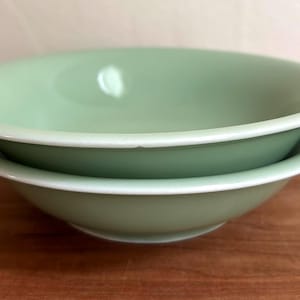 May include: Two stacked round bowls in a solid light sage green colour. The bowls feature a smooth, glossy finish with a simple, wide rim design. The bowls are identical in shape and size, creating a uniform appearance when stacked together.