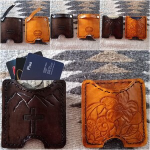 May include: A collection of square leather card holders in dark brown and tan, featuring hand-tooled designs including a mountain range with a cross and floral patterns. Some holders include a leather strap with a metal snap closure.