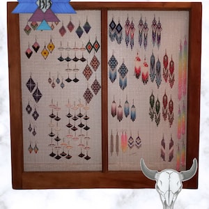 May include: A wooden display case with a wire mesh backing holds a variety of colourful beaded earrings. The earrings are arranged in rows and feature geometric patterns and fringe details. The display case is decorated with a skull and the text "Buckskin Beading PhotoRoom".