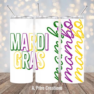 May include: Three white insulated tumblers with silver straws. The first tumbler has the words "Mardi Gras" in green, purple, and yellow. The second tumbler has the word "mambo" in green, purple, and yellow. The third tumbler has the word "mambo" in green, purple, and yellow.