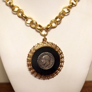 Puede incluir: Un collar de cadena dorada con un colgante redondo con fondo negro y una moneda de plata con el perfil de un hombre.