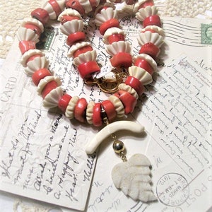 May include: A coral and white bead necklace with a carved bone pendant in the shape of a leaf. The necklace is made of coral and bone beads and has a gold clasp.