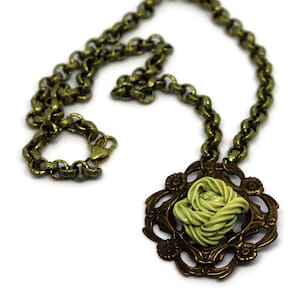 Puede incluir: Un collar de cadena dorada con un colgante que presenta una rosa de tela verde retorcida en un marco de filigrana dorada.