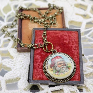 Puede incluir: Un collar de estilo vintage con un colgante redondo que presenta una ilustración de Santa Claus. El colgante está rodeado por un marco dorado con el texto "Markeson's Toy Department".