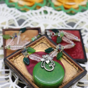 May include: A green pendant necklace with a silver floral design, displayed on a vintage frame. The necklace is surrounded by other jewelry pieces with green and clear beads. The background includes lace and decorative flowers.