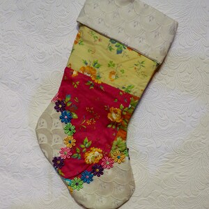 May include: A patchwork Christmas stocking with a cream-colored cuff, featuring floral patterns in shades of yellow, pink, and red. The stocking is adorned with colorful beaded flowers.