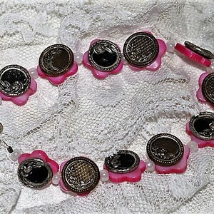 May include: A pink and silver necklace with flower-shaped charms. Each charm has a silver center with a floral design. The necklace is strung on a white cord with small white beads.