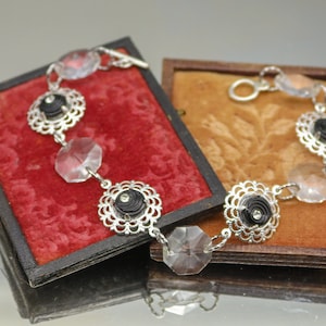 May include: A silver bracelet with black and white accents. The bracelet features a series of clear crystal beads and black rosettes with silver trim. The bracelet is displayed on a red velvet box.