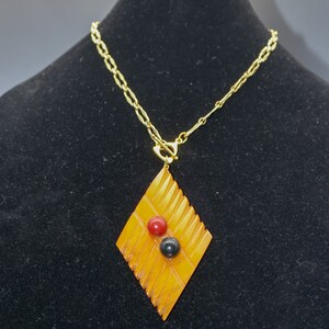Op de afbeelding: Een gouden ketting met een ruitvormige hanger van barnsteen met drie ronde kralen in rood en zwart.