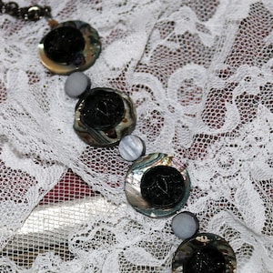 May include: A necklace with a silver chain and multiple round iridescent shell buttons with black centers. The necklace is on a white lace background.