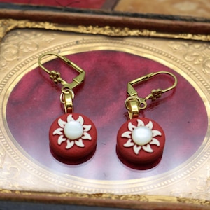 Könnte beinhalten: Ein Paar Ohrringe mit einem roten kreisförmigen Design und einem weißen Blumenmuster. Die Ohrringe haben goldfarbene Haken. Die Ohrringe werden in einem dekorativen Rahmen mit rotem Hintergrund präsentiert.