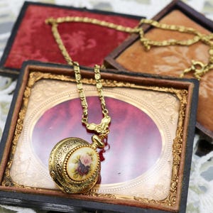 Può includere: Una collana con orologio da tasca color oro con un design floreale, appoggiata su un espositore decorativo. L'orologio presenta un design dettagliato e ornato con una catena. Lo sfondo include due scatole espositive foderate in velluto.