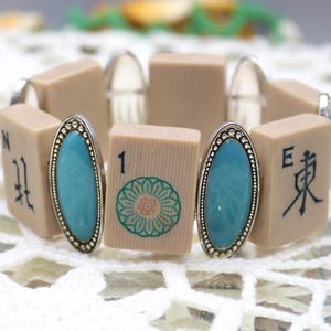 Könnte beinhalten: Ein Armband aus beigefarbenen Mahjong-Steinen mit ovalen türkisfarbenen Akzenten. Das Armband hat silberfarbenes Metall und ist mit schwarzen chinesischen Schriftzeichen und einem grün-orangen Blumenmuster verziert.