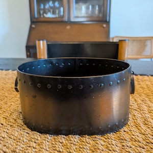 Puede incluir: Un recipiente redondo de metal oscuro con detalles remachados alrededor del borde y pequeñas asas en los laterales. El recipiente tiene un acabado ligeramente desgastado, lo que sugiere una estética antigua. Se encuentra sobre una estera tejida de tono natural.