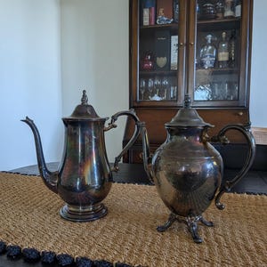 May include: Two antique silver-plated teapots on a woven brown table runner. The teapot on the left has a tall, slender body and a curved spout. The teapot on the right is rounder and sits on three feet. A dark wooden cabinet is in the background.